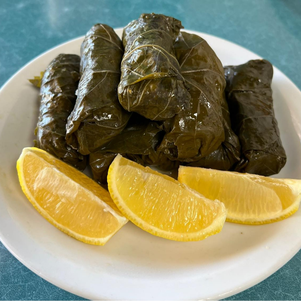 A white plate holds several dark green dolmades, or stuffed grape leaves, with three lemon wedges.